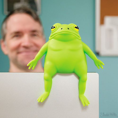 A Splat Frog, bright green and filled with sand, rests on a white surface while a smiling man is blurred in the background.