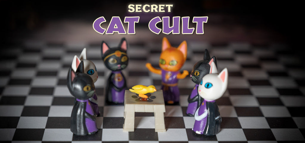 Vinyl cats in robes are seated around a table with a rubber duck. Above them, the text reads "Secret Cat Cult," suggesting their mysterious feline society.